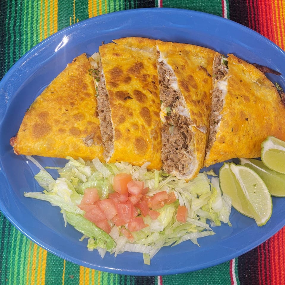 A large, golden-brown quesadilla, sliced into four pieces, filled with meat and cheese, served with lettuce, diced tomatoes, and lime wedges on a blue plate.