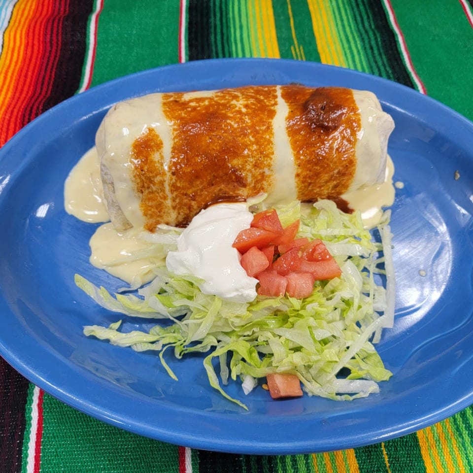 A large, golden-brown burrito covered in white sauce, served with shredded lettuce, diced tomatoes, and sour cream on a blue plate.
